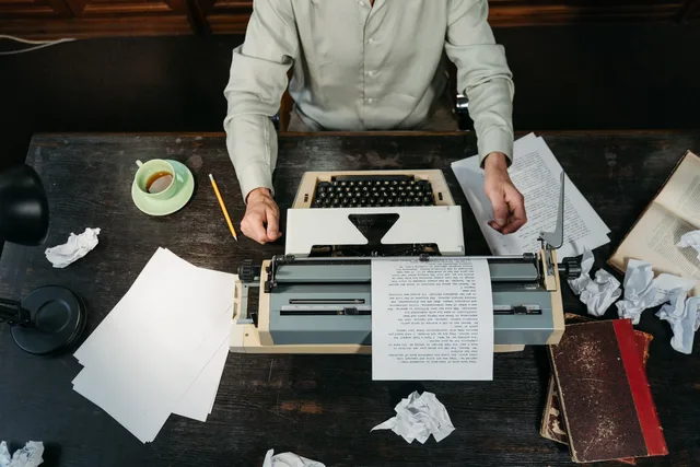 An overhead view of a workspace with a vintage typewriter, scattered papers, and a person writing. Sponsored article production